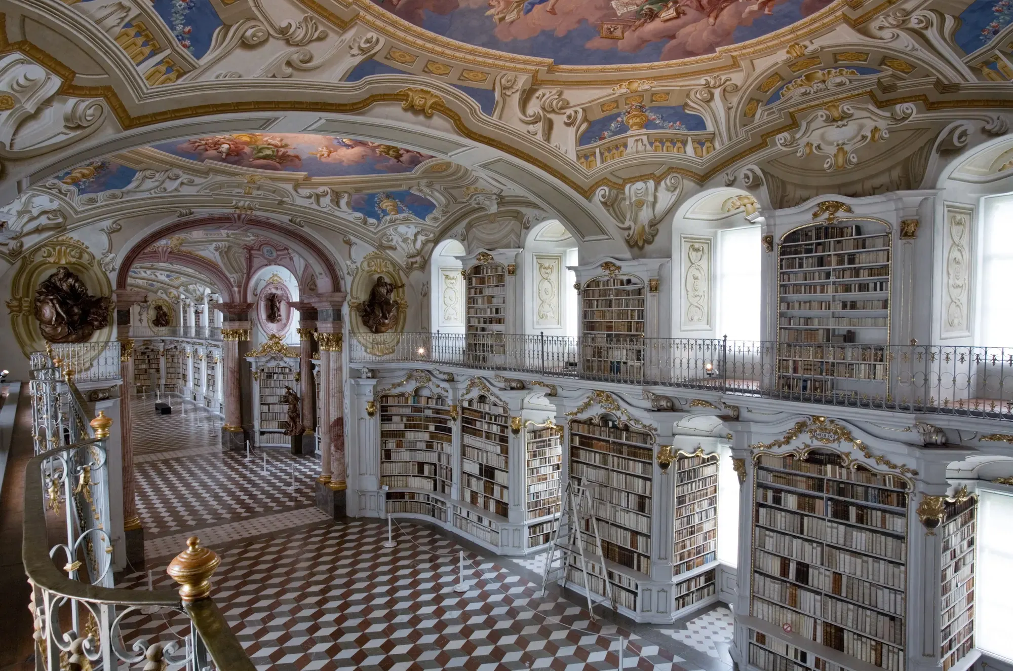 Baroque library hall with white and gold bookcases, painted ceiling frescoes, a railed gallery and a chequered marble floor
