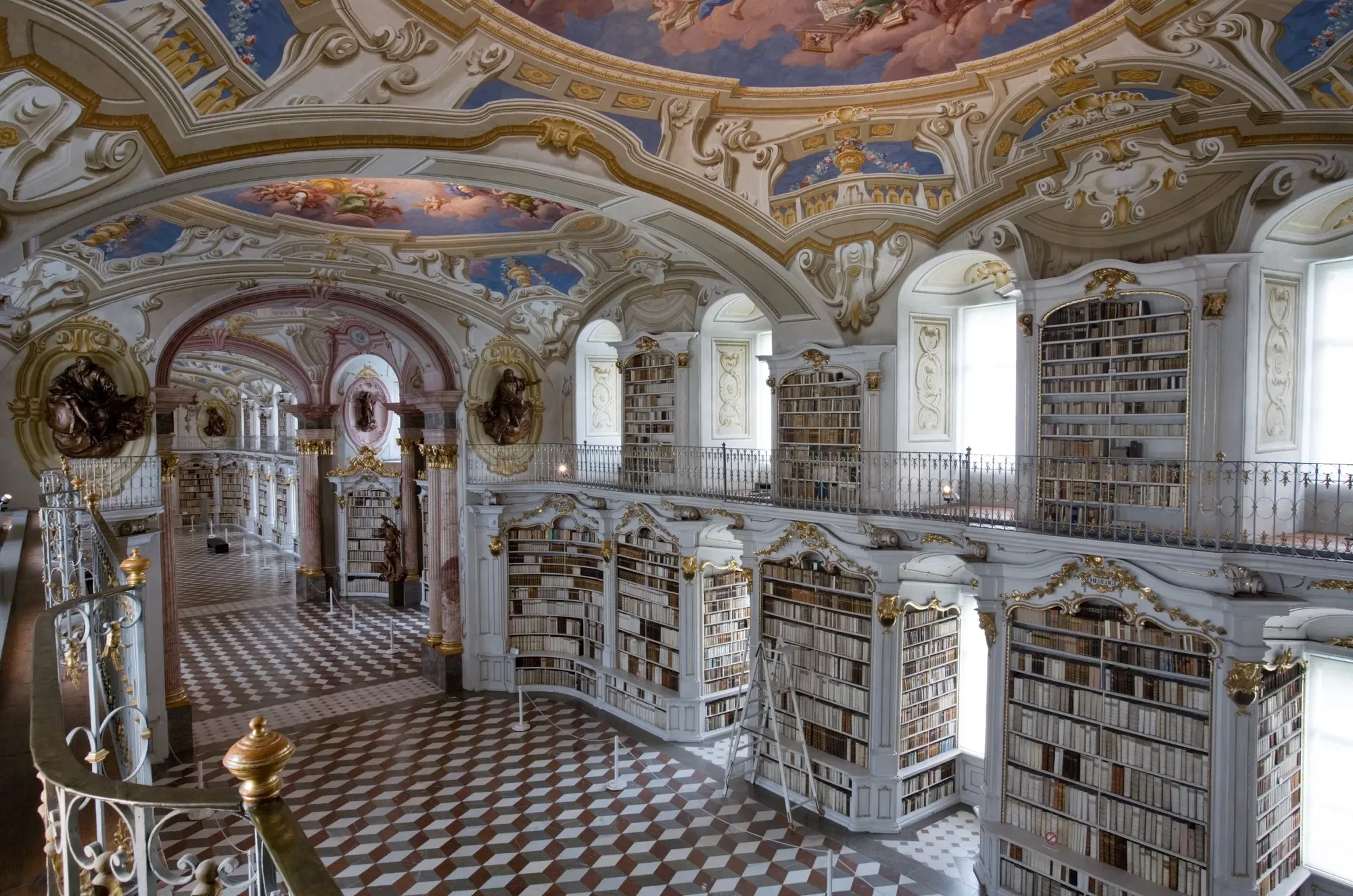 Baroque library hall with white and gold bookcases, painted ceiling frescoes, a railed gallery and a chequered marble floor