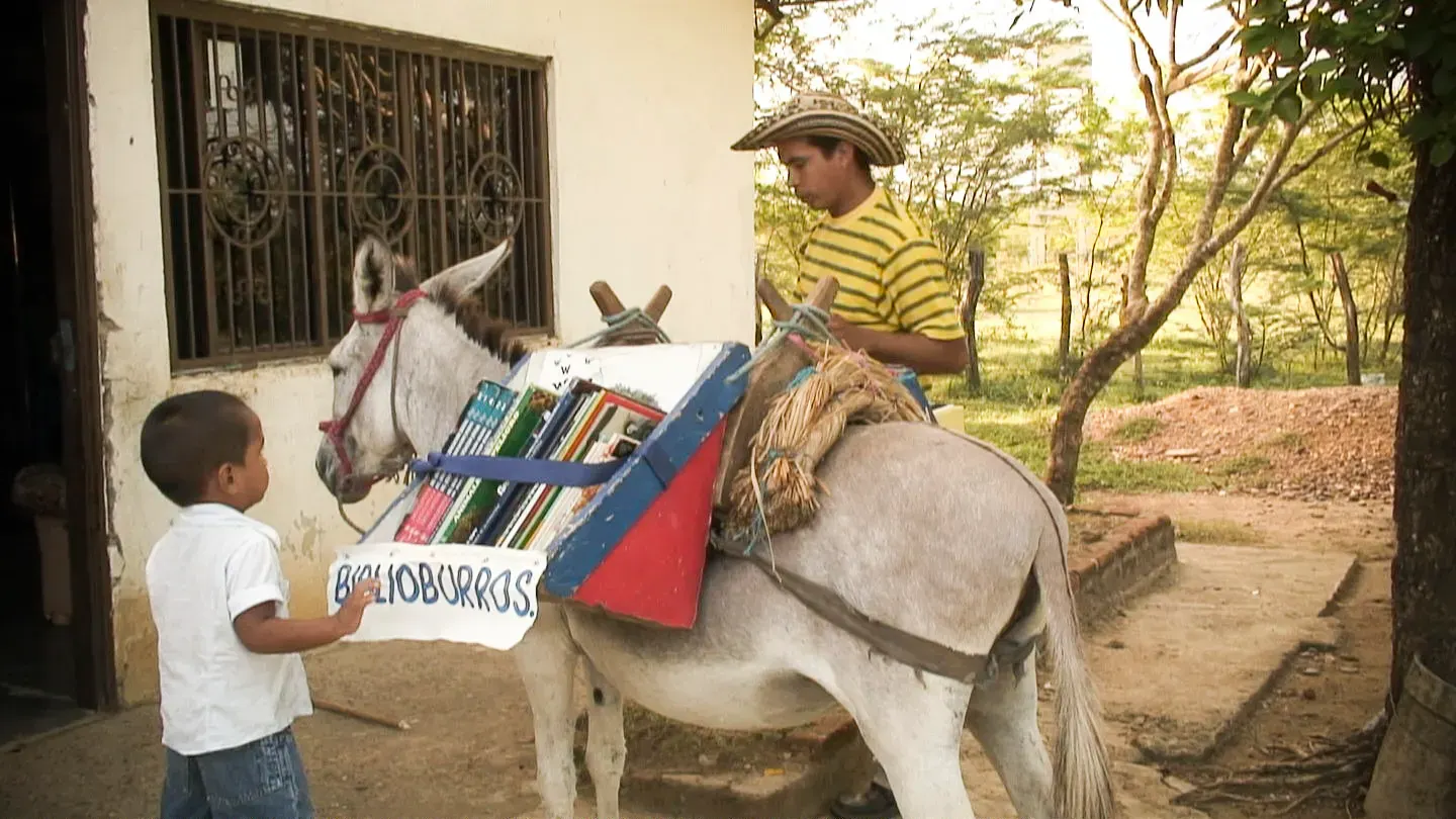 A donkey carrying books under a 'Biblioburro' sign, while a man and a child choose a book