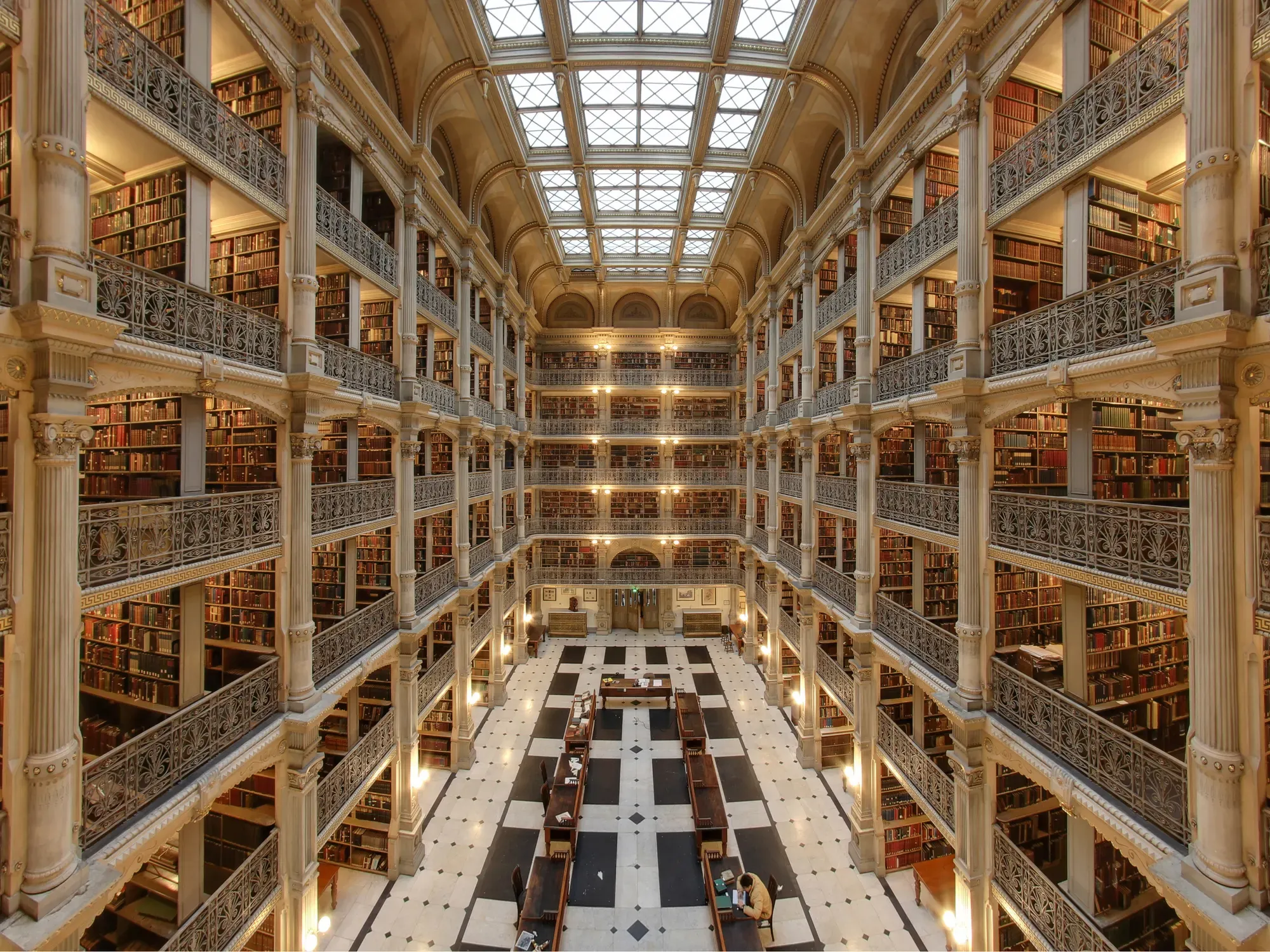 The atrium of the George Peabody Library: five tiers of cast-iron balconies lined with books rise to a skylight above reading tables