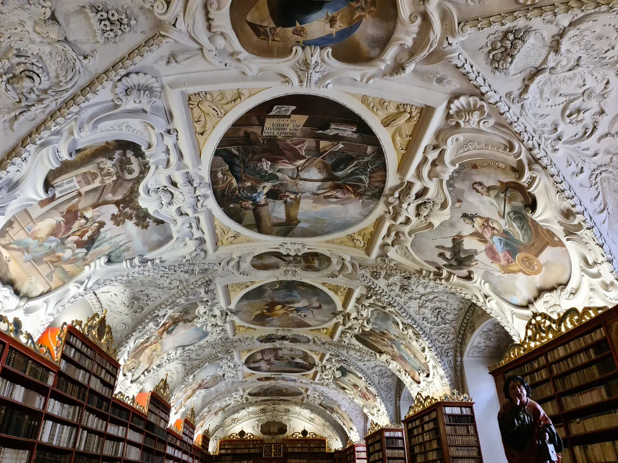 A baroque library hall at Strahov, its white stucco ceiling framing painted frescoes in cartouches above rows of bookcases
